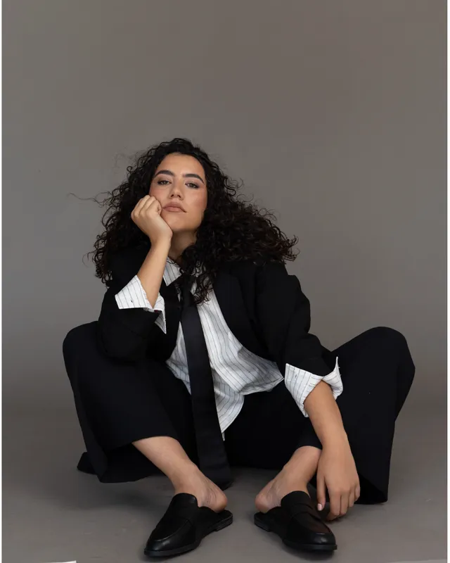 Aurora in a black suit and tie, sitting, hand on her chin